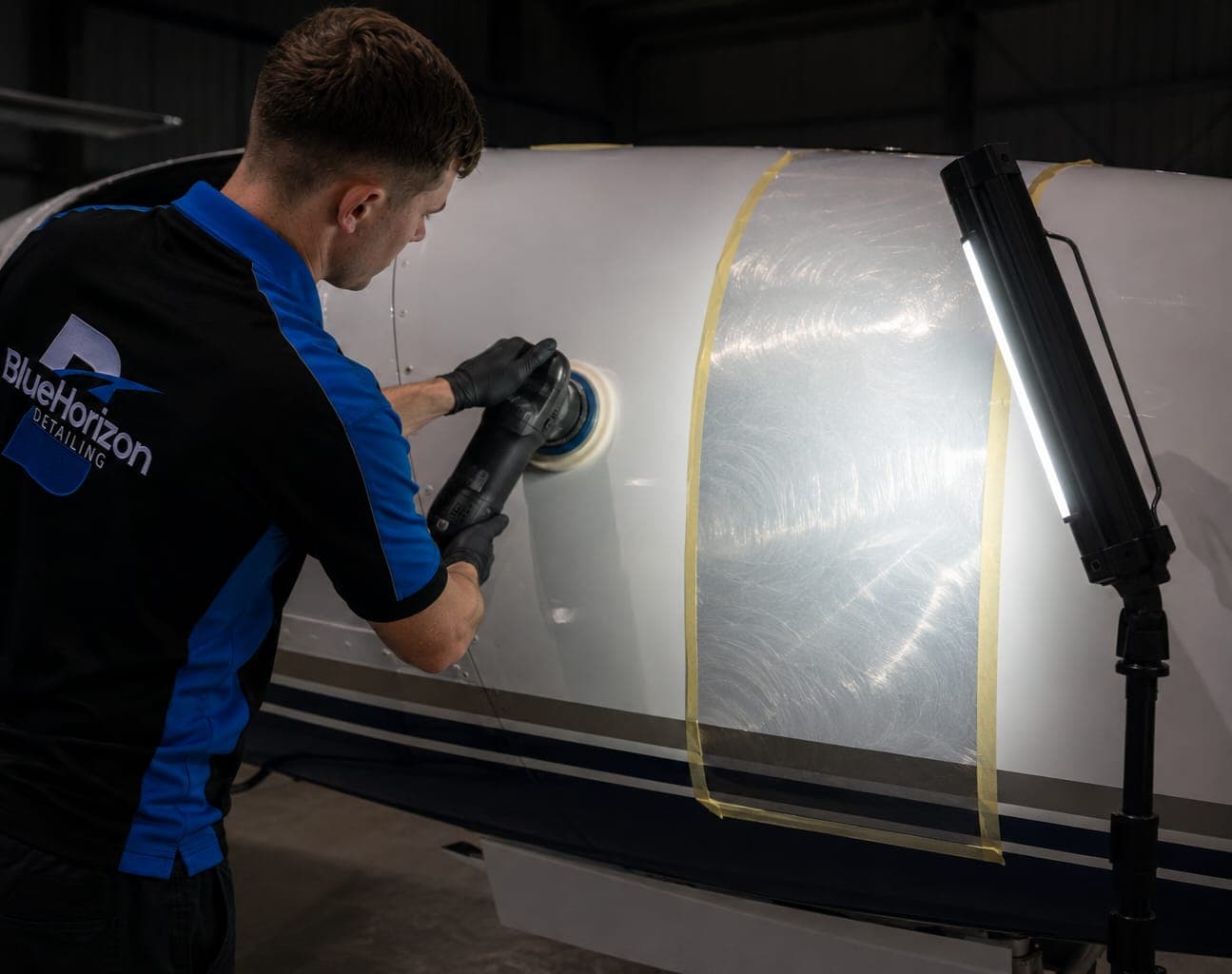 A Blue Horizon technician machine-polishing an aircraft fuselage panel, swirl marks revealed under an inspection lamp